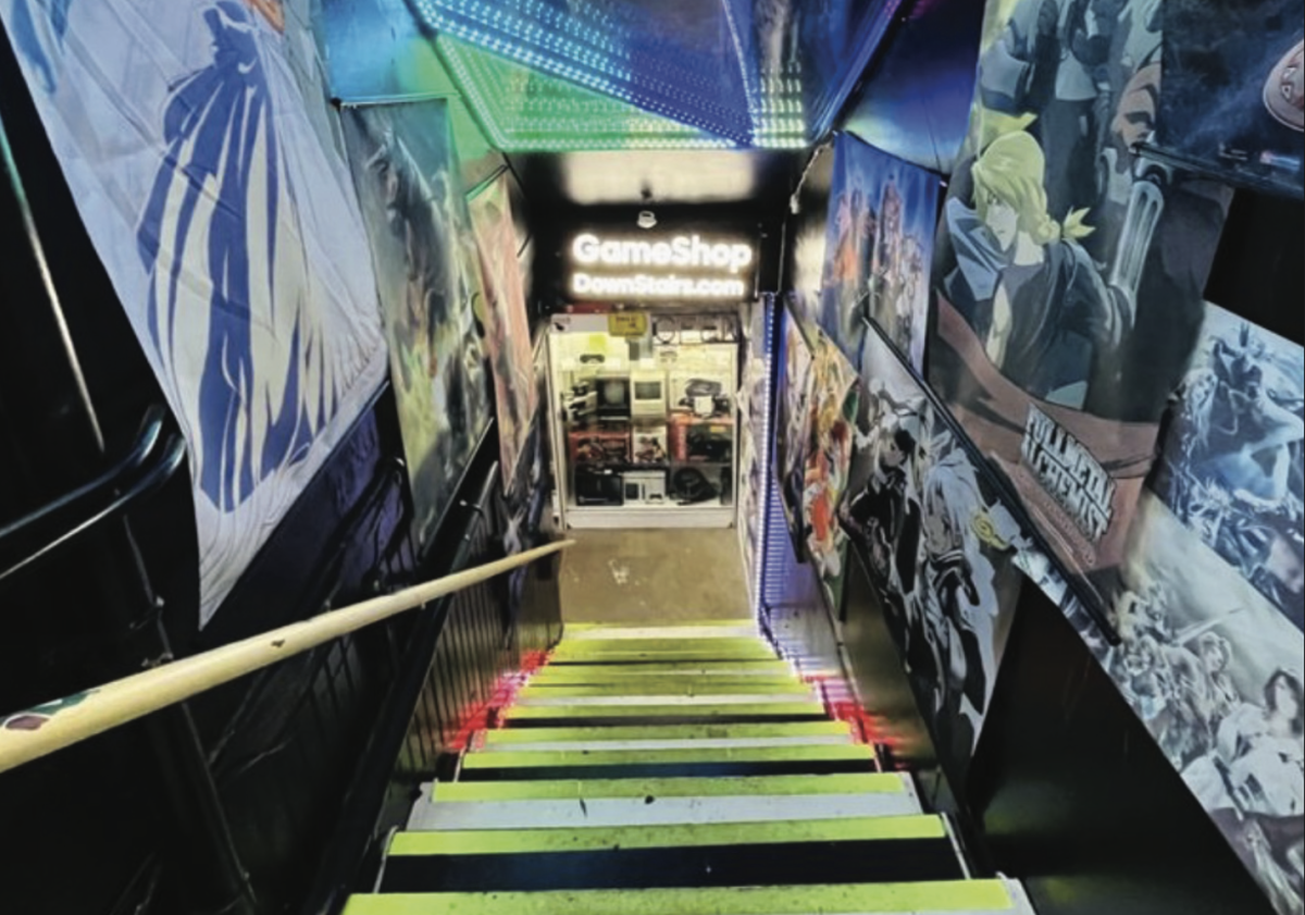 Colorful lights flash overhead against a corridor of anime posters descending into the Gameshop Downstairs entrance.